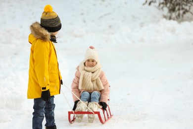 Photo of Little boy pulling sledge with his sister through snow in winter park, space for text