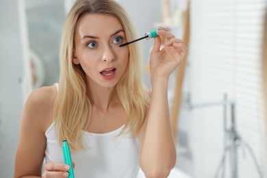 Beautiful woman applying mascara near mirror in bathroom