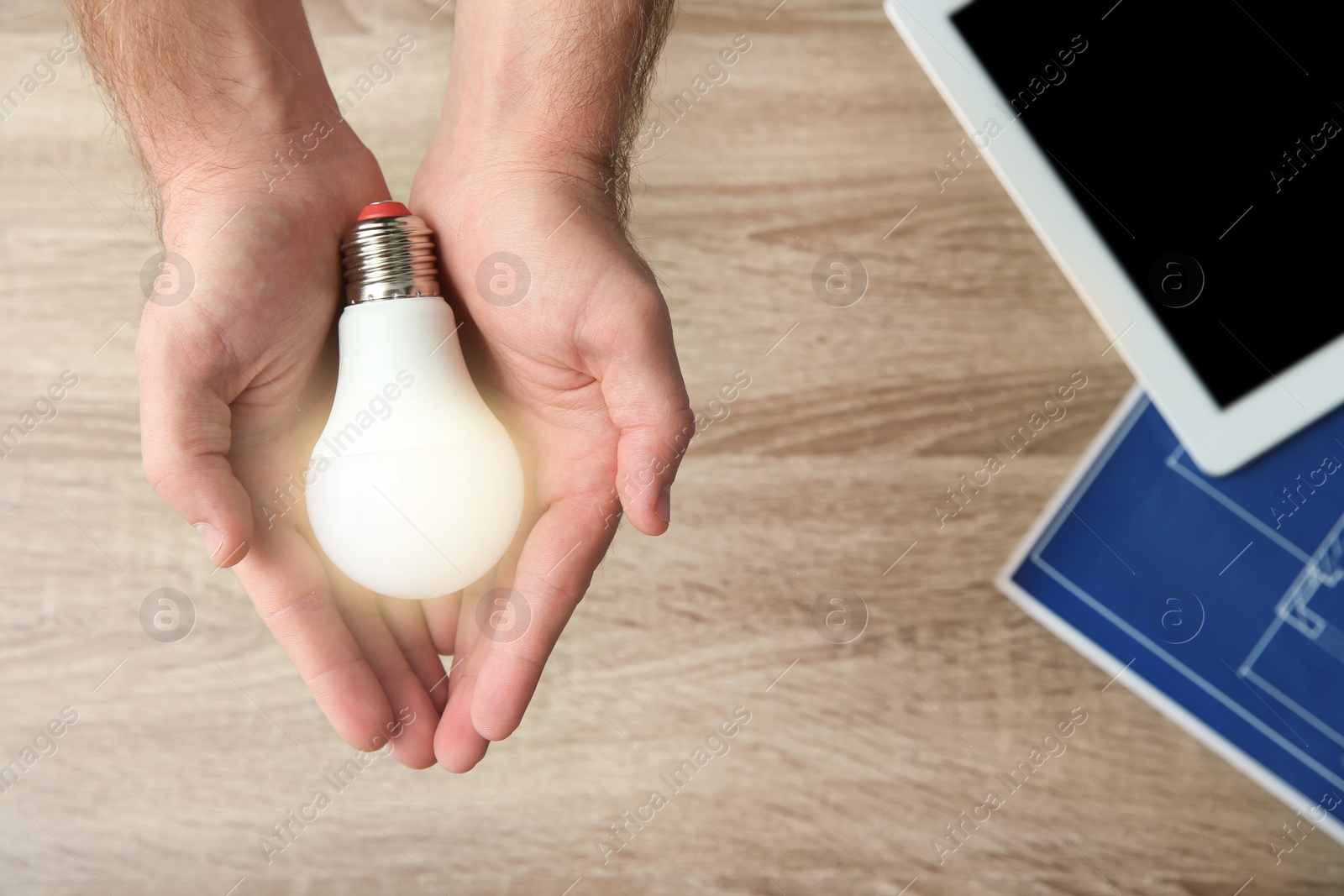 Photo of Man holding lamp bulb over table with tablet and blueprint, top view. Space for text