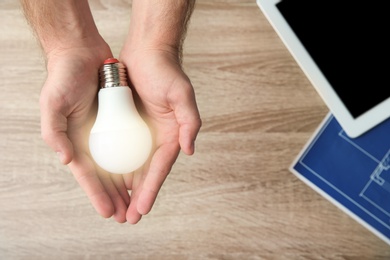 Photo of Man holding lamp bulb over table with tablet and blueprint, top view. Space for text