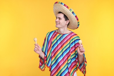 Young man in Mexican sombrero hat and poncho with maracas on yellow background. Space for text