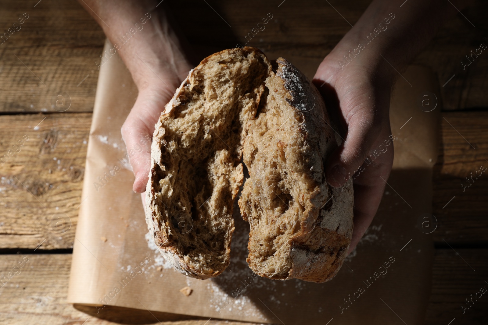 Photo of Man breaking loaf of fresh bread at wooden table, top view