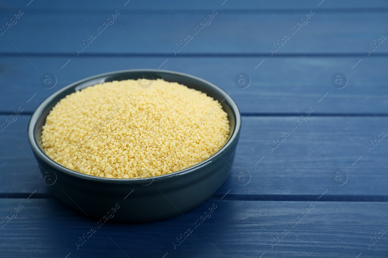 Photo of Bowl of raw couscous on blue wooden table, closeup. Space for text
