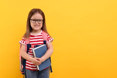 Photo of Cute little girl in glasses with book and backpack on orange background. Space for text