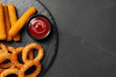 Photo of Tasty ketchup, onion rings and cheese sticks on dark textured table, top view. Space for text