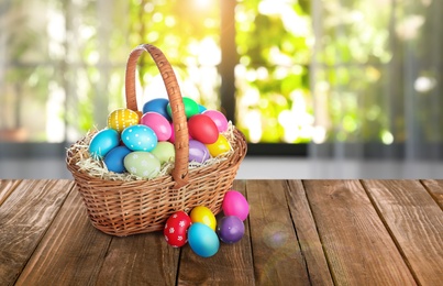 Image of Wicker basket with bright painted Easter eggs on wooden table, space for text