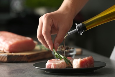 Photo of Man cooking fresh raw meat with oil and rosemary on table against blurred background, closeup