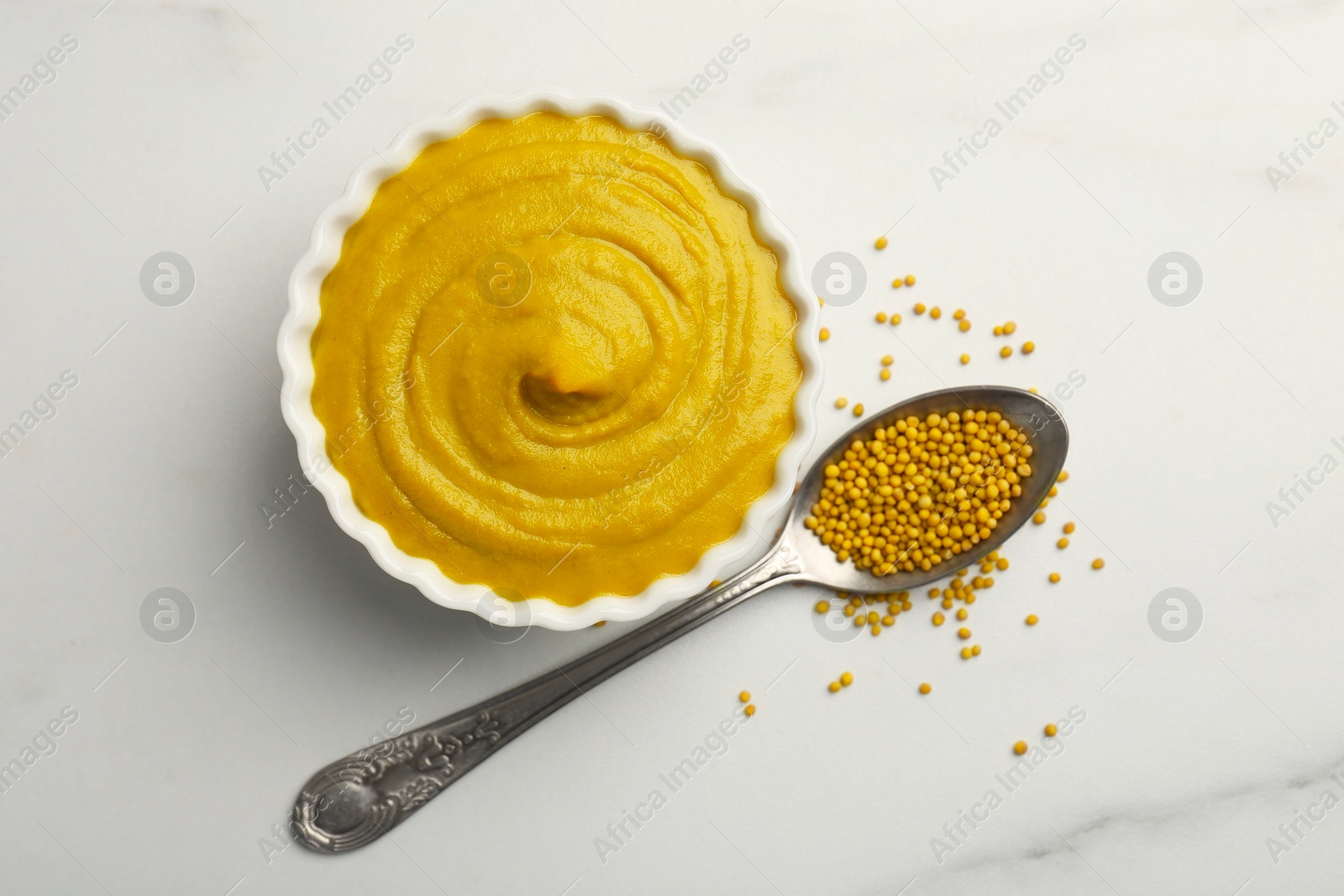 Photo of Tasty mustard sauce in bowl and spoon with dry seeds on white marble table, flat lay