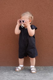 Photo of Cute little girl wearing stylish clothes with sunglasses and pacifier near brown wall outdoors