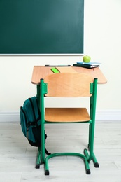 Photo of Wooden school desk with stationery, apple and backpack near chalkboard in classroom