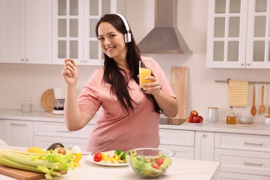 Photo of Happy overweight woman with headphones and glass of juice dancing near table in kitchen. Healthy diet