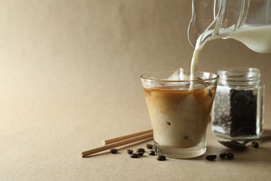 Photo of Pouring milk into glass with refreshing iced coffee on beige background, closeup. Space for text