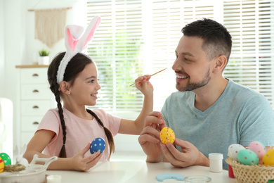 Happy daughter with bunny ears headband and her father having fun while painting Easter eggs at home