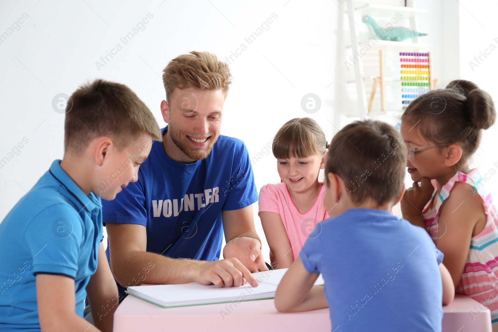 Photo of Young volunteer reading book with children at table indoors