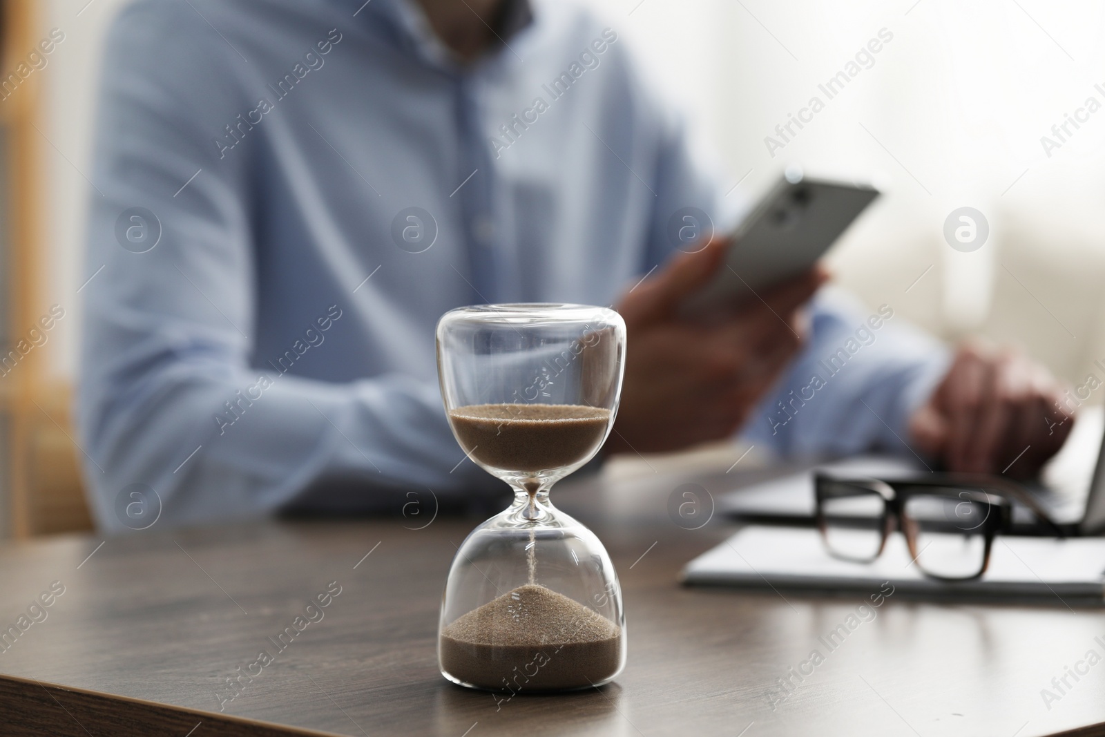 Photo of Hourglass with flowing sand on desk. Man using smartphone while working on laptop indoors, selective focus