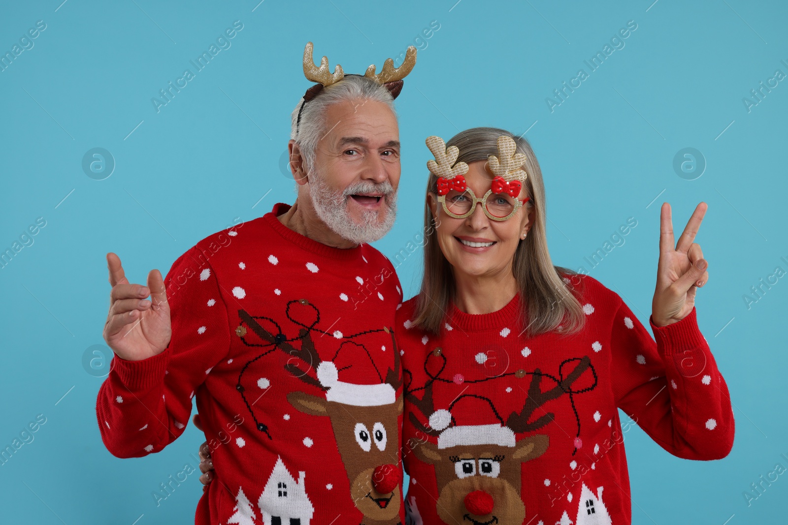 Photo of Senior couple in Christmas sweaters, reindeer headband and funny glasses on light blue background