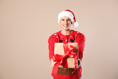 Happy man in Christmas sweater and Santa hat holding gift box on beige background