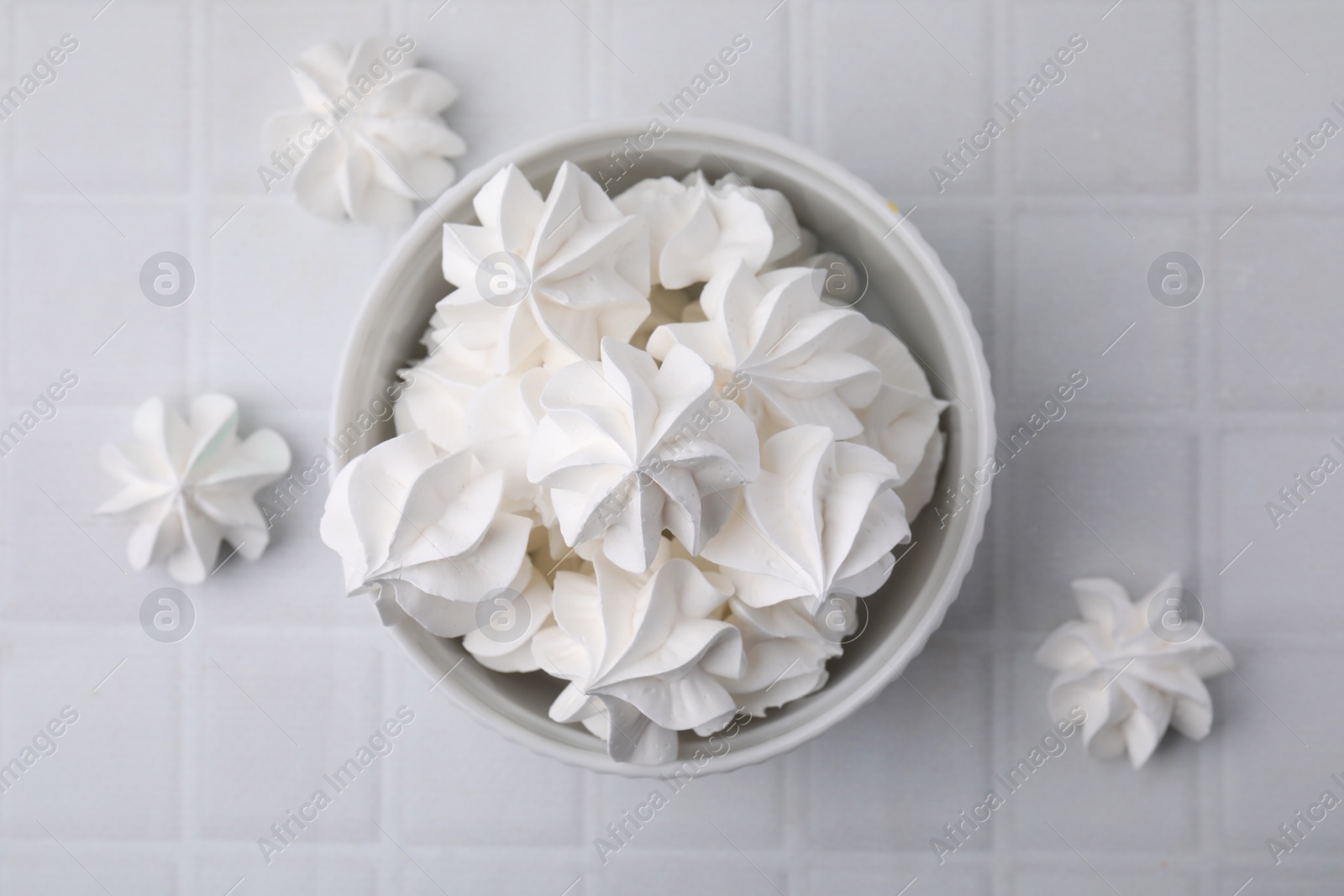 Photo of Delicious meringue cookies in bowl on white tiled table, flat lay