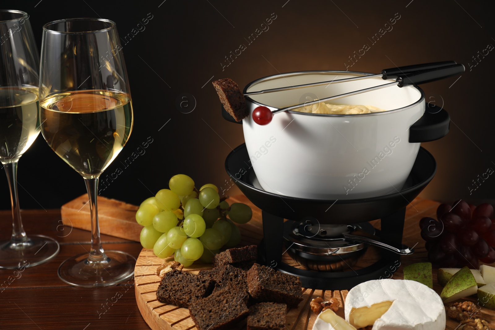 Photo of Forks with pieces of grape, bread, fondue pot with melted cheese, wine and snacks on wooden table, closeup