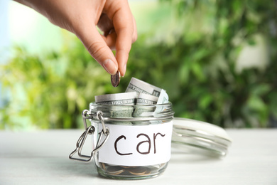 Photo of Woman putting coin into jar with tag CAR on white wooden table, closeup