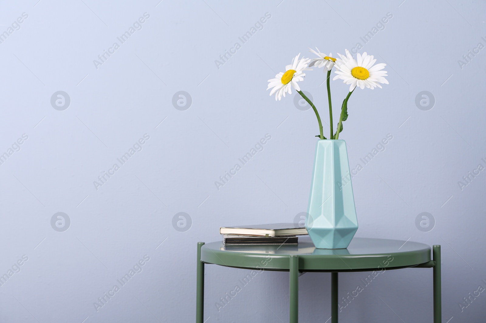 Photo of Beautiful tender chamomile flowers in vase and notebooks on table against light background, space for text