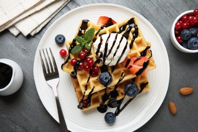 Photo of Delicious Belgian waffles with ice cream, berries and chocolate sauce served on grey textured table, flat lay
