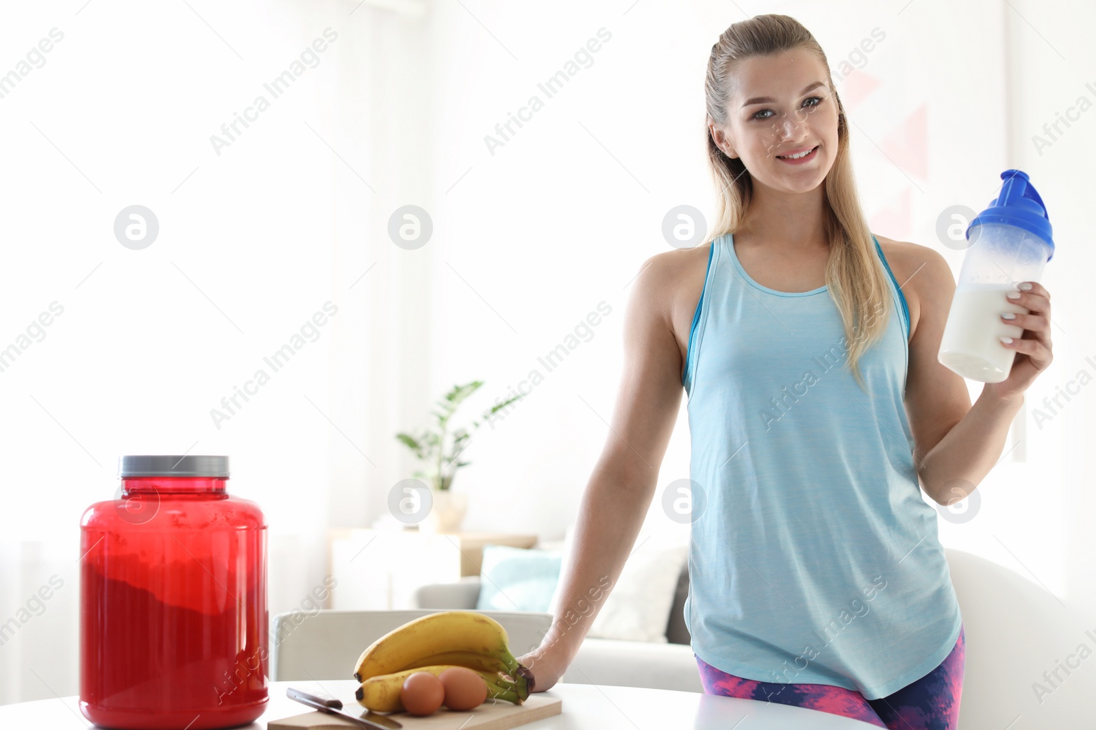 Photo of Young woman holding bottle of protein shake near table with ingredients in room. Space for text
