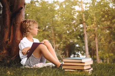 Cute little boy reading book on green grass near tree in park
