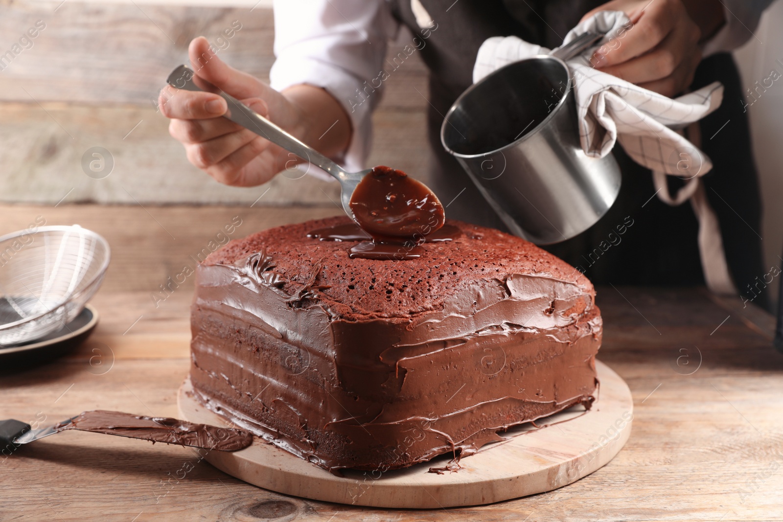 Photo of Woman pouring chocolate cream into homemade sponge cake at wooden table, closeup