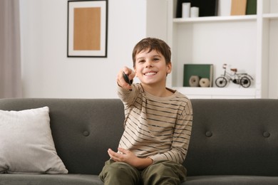 Photo of Happy boy changing TV channels with remote control on sofa at home