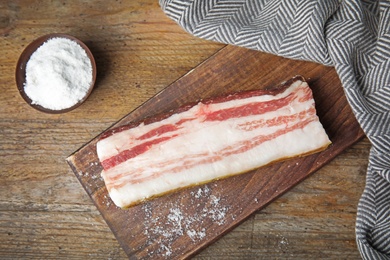 Photo of Board with bacon and salt on wooden background, flat lay