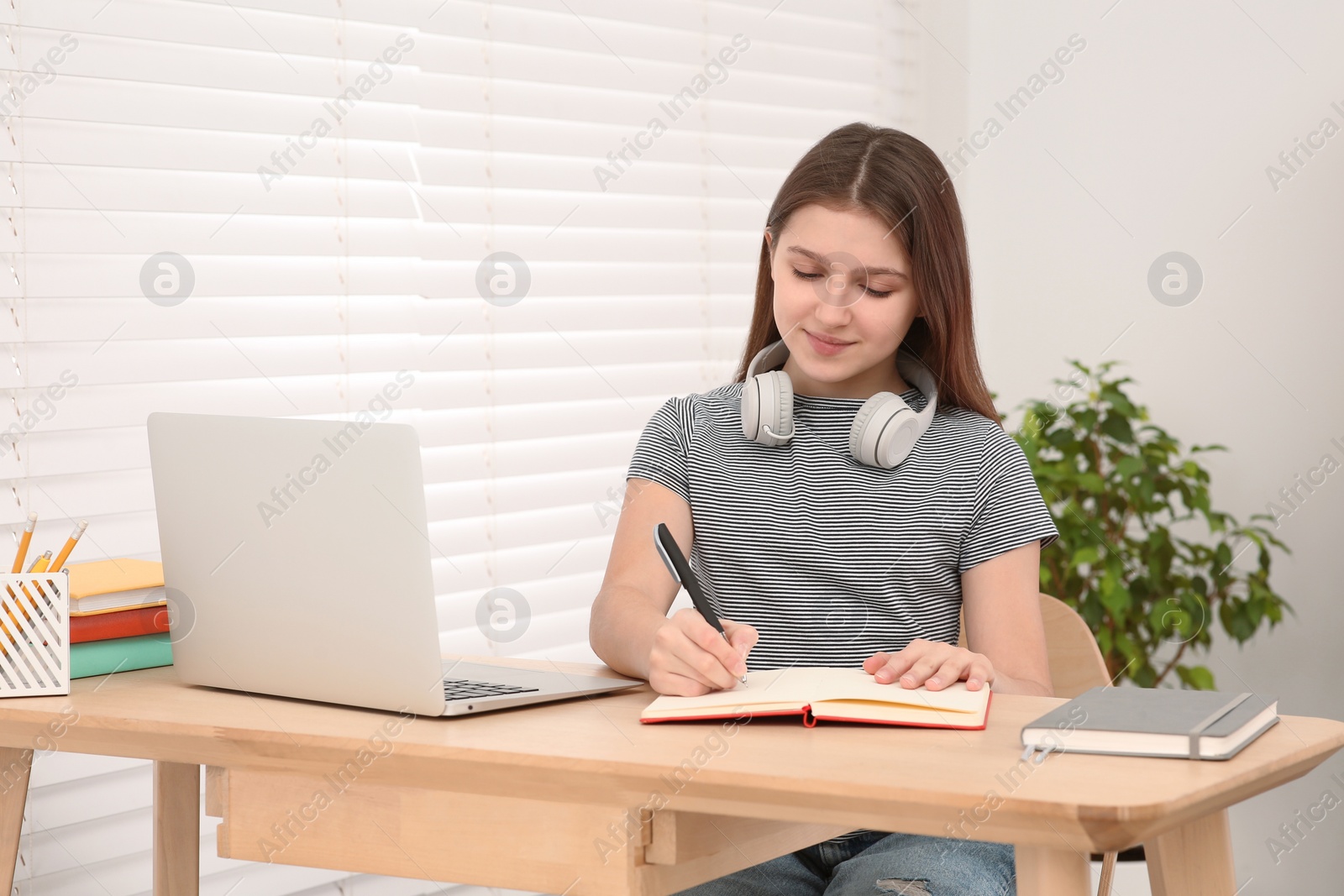 Photo of Cute girl with headphones writing in notepad near laptop at desk in room. Home workplace