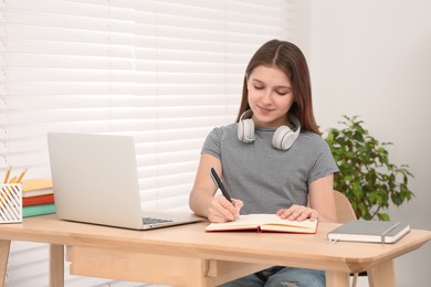 Photo of Cute girl with headphones writing in notepad near laptop at desk in room. Home workplace