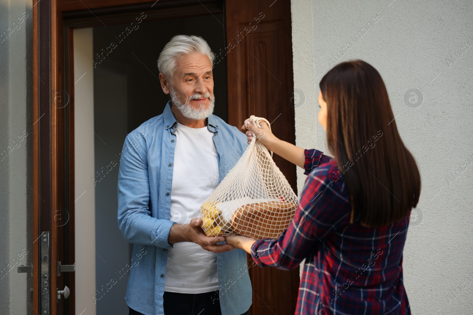 Photo of Helping neighbours. Young woman with net bag of products visiting senior man outdoors