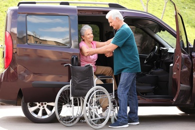 Photo of Mature man helping senior woman to get out from van into wheelchair outdoors