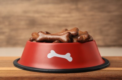 Photo of Red bowl with bone shaped dog cookies on wooden board, closeup