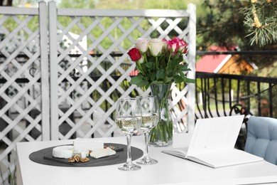 Photo of Vase with roses, open book, glasses of wine and snacks on white table at balcony