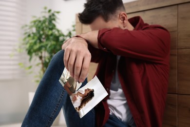 Photo of Crying man sitting on floor indoors, focus on torn photo. Divorce concept