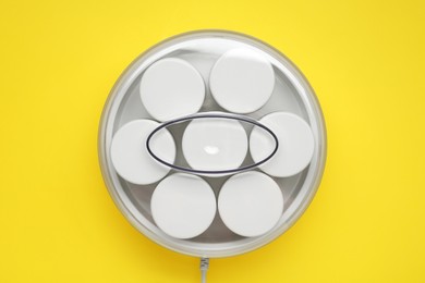 Modern yogurt maker with jars on yellow background, top view