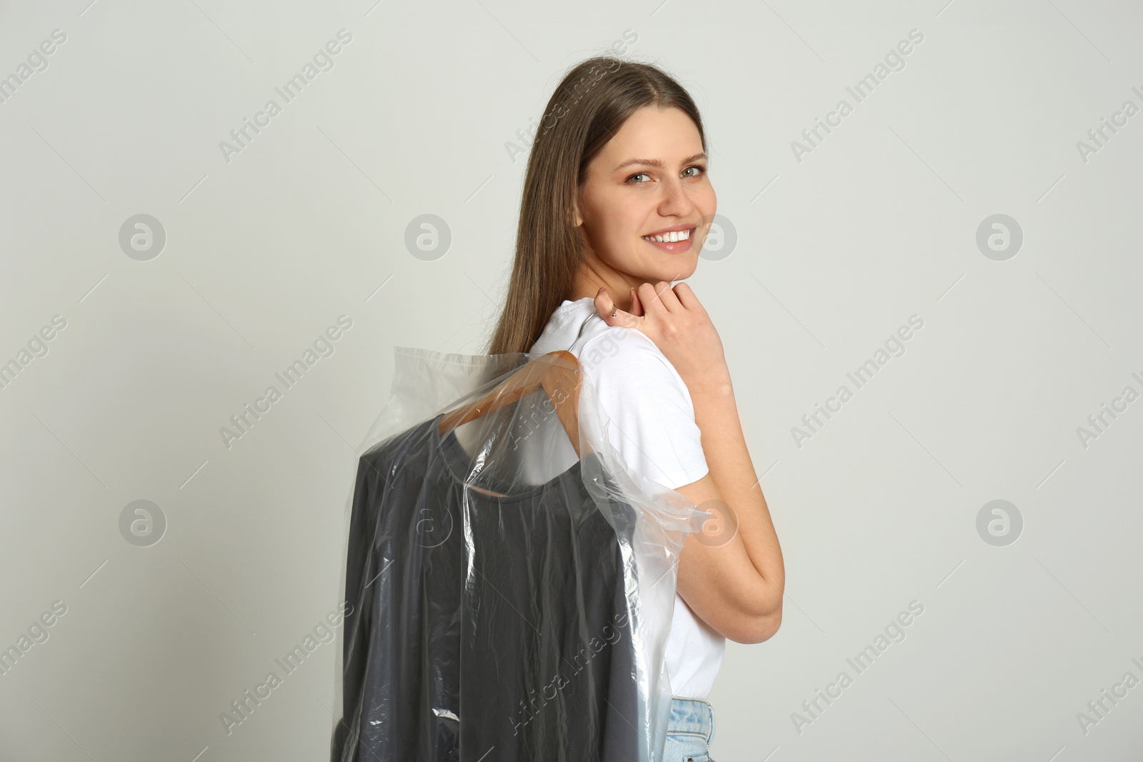 Photo of Young woman holding hanger with dress in plastic bag on light grey background. Dry-cleaning service