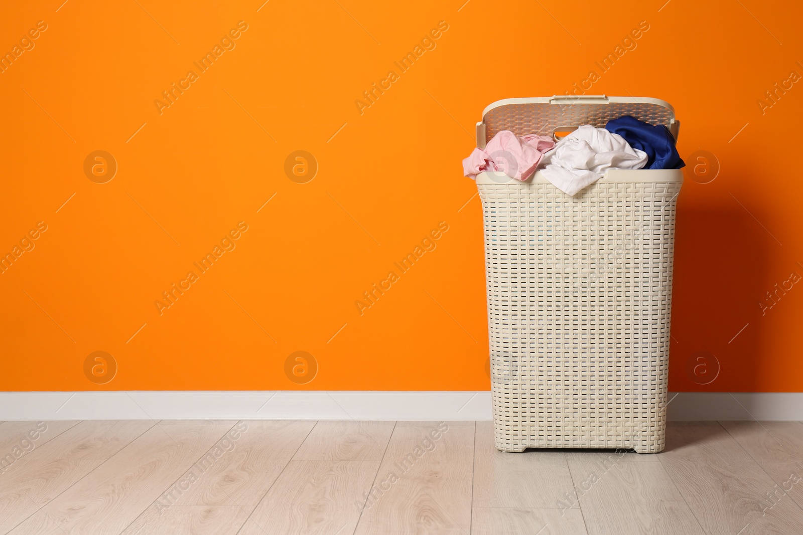 Photo of Open laundry basket with clothes near orange wall indoors, space for text