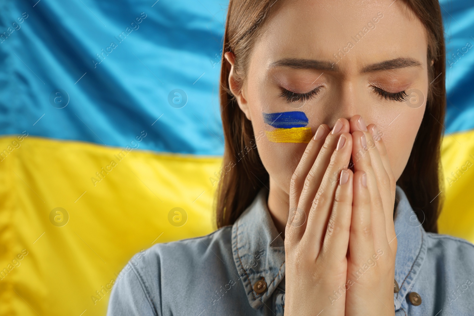 Photo of Sad young woman with clasped hands near Ukrainian flag, closeup. Space for text