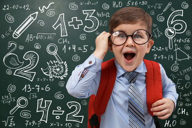 Image of Funny little child wearing glasses near chalkboard with mathematical equations. First time at school