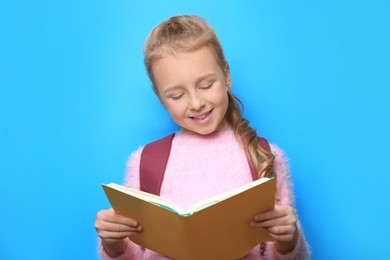 Photo of Happy little girl with backpack reading book on light blue background