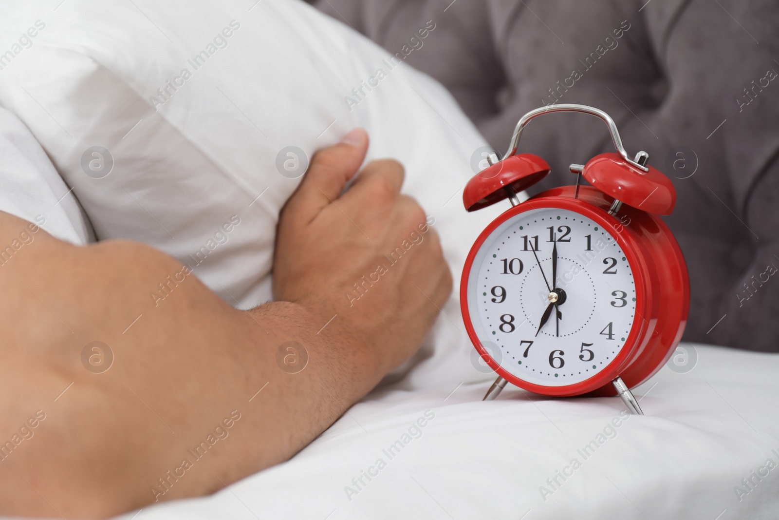 Photo of Alarm clock and man covering head with pillow in bedroom. Time of day