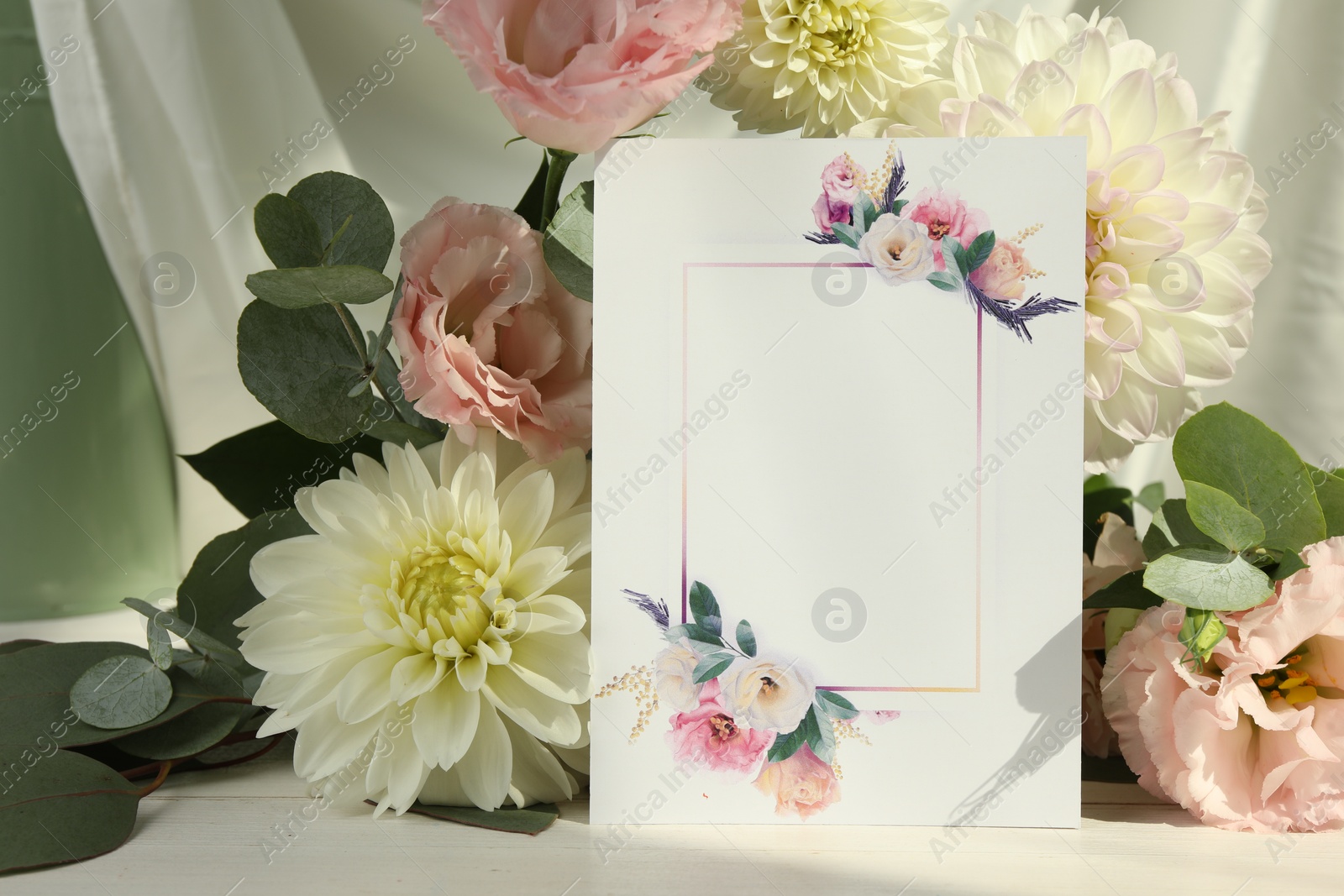 Photo of Blank invitation card, eucalyptus leaves and flowers on white wooden table. Space for text