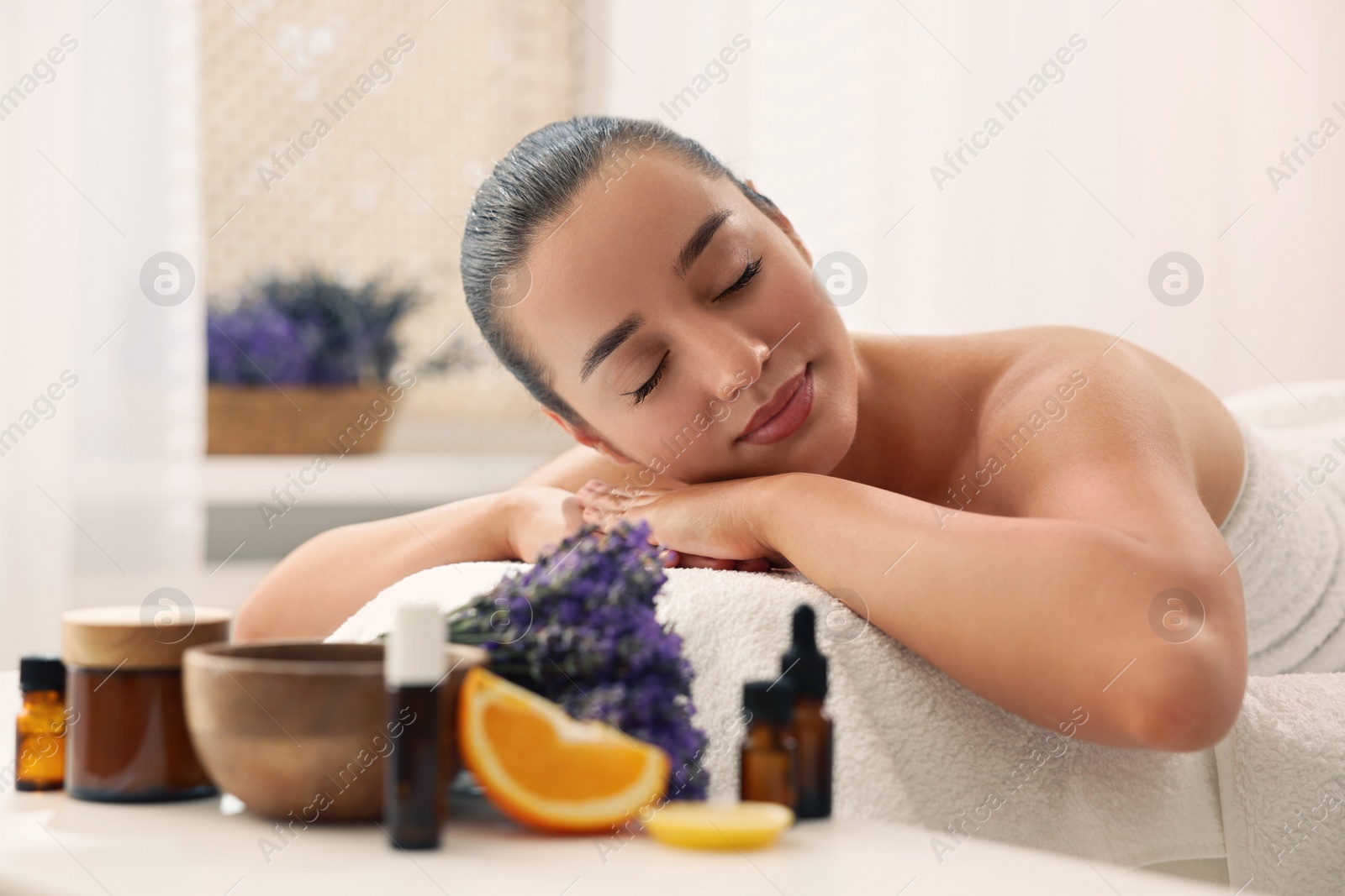 Photo of Beautiful young woman relaxing on massage couch and bottles of essential oil with ingredients on table in spa salon