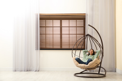 Young woman relaxing in hanging chair near window at home. Space for text