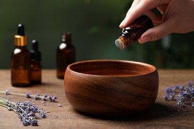 Photo of Woman dripping essential oil from bottle into bowl near lavender at wooden table, closeup. Space for text