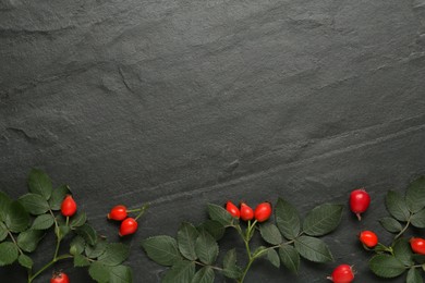 Photo of Ripe rose hip berries with green leaves on black table, flat lay. Space for text